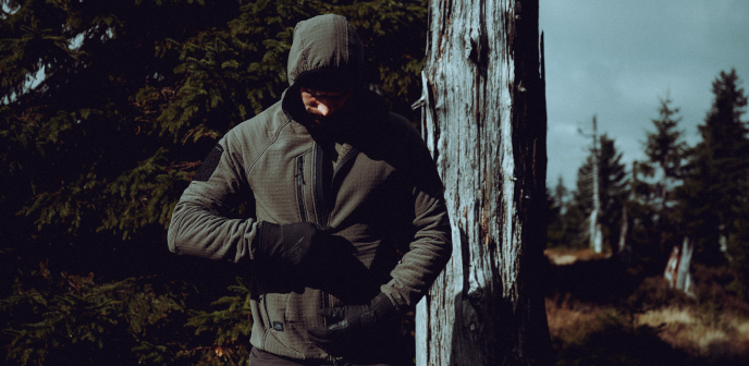 Zimowe działanie w terenie &ndash; mężczyzna siedzi na ośnieżonym pniu w lesie, ubrany w ciepłą odzież outdoorową